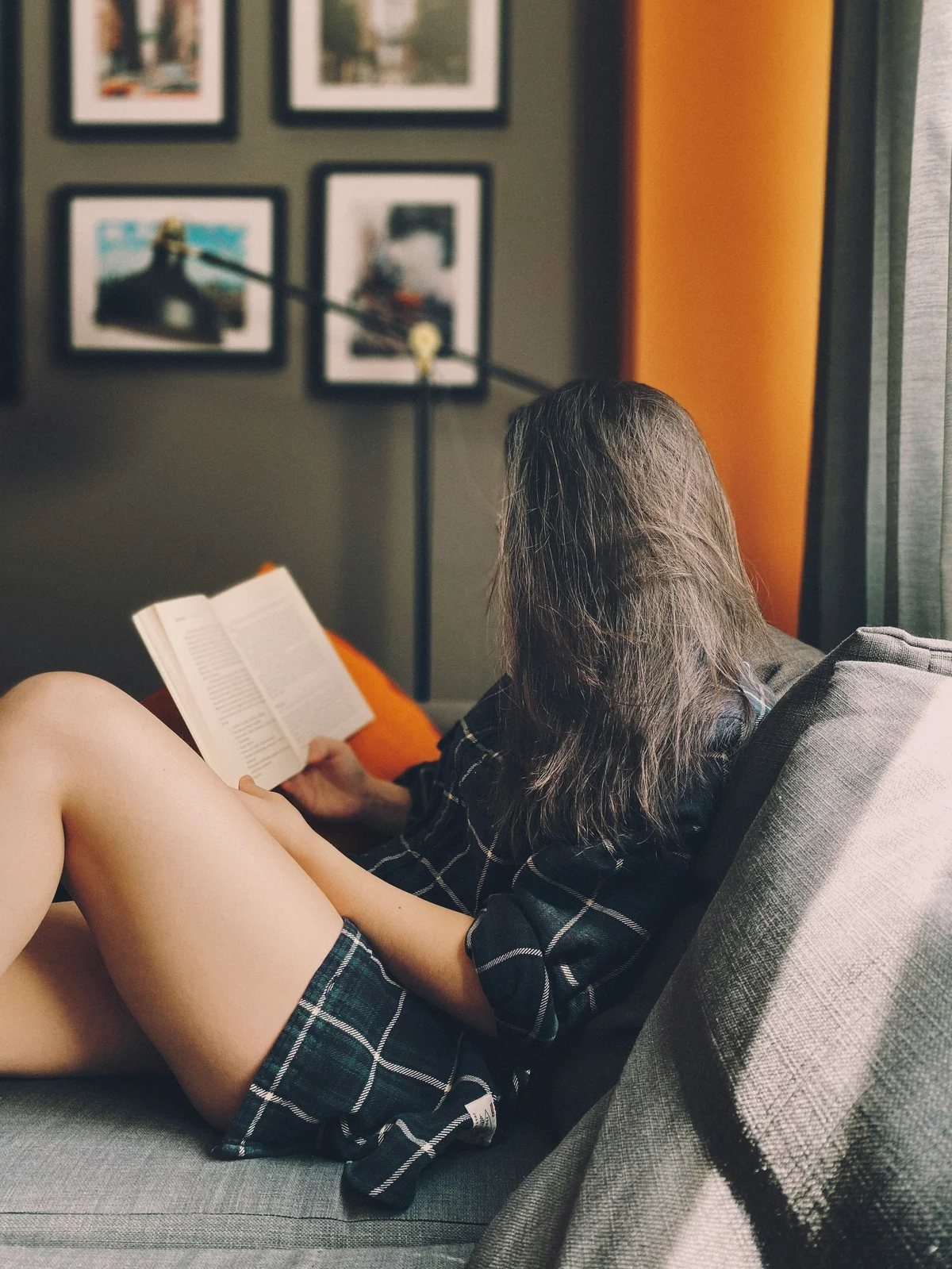 Vue de profil d'une femme assise sur un canapé en train de lire un livre, ses longs cheveux masquant son visage ; elle porte un short en coton ample et une chemise à carreaux vert foncé