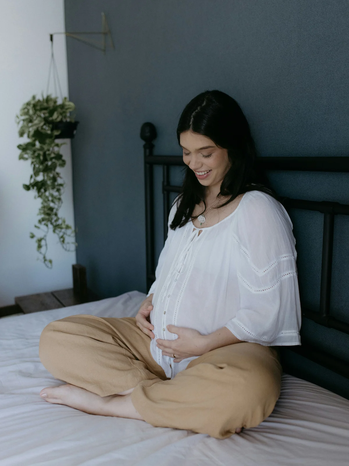 Une femme enceinte assise sur un lit, jambes croisées, en chemise blanche et pantalon beige, les mains sur son ventre, souriant vers le bas