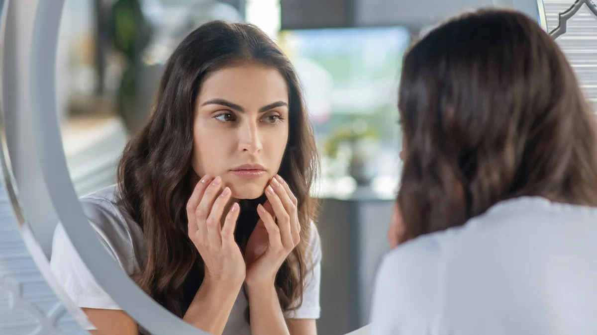 Une femme aux longs cheveux bruns et aux sourcils nets et bien définis se regarde dans le miroir, les mains délicatement posées sur son visage
