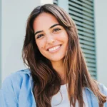 Une femme aux longs cheveux bruns et aux sourcils bien définis se tient à l'extérieur, souriante, devant un bâtiment aux volets verts