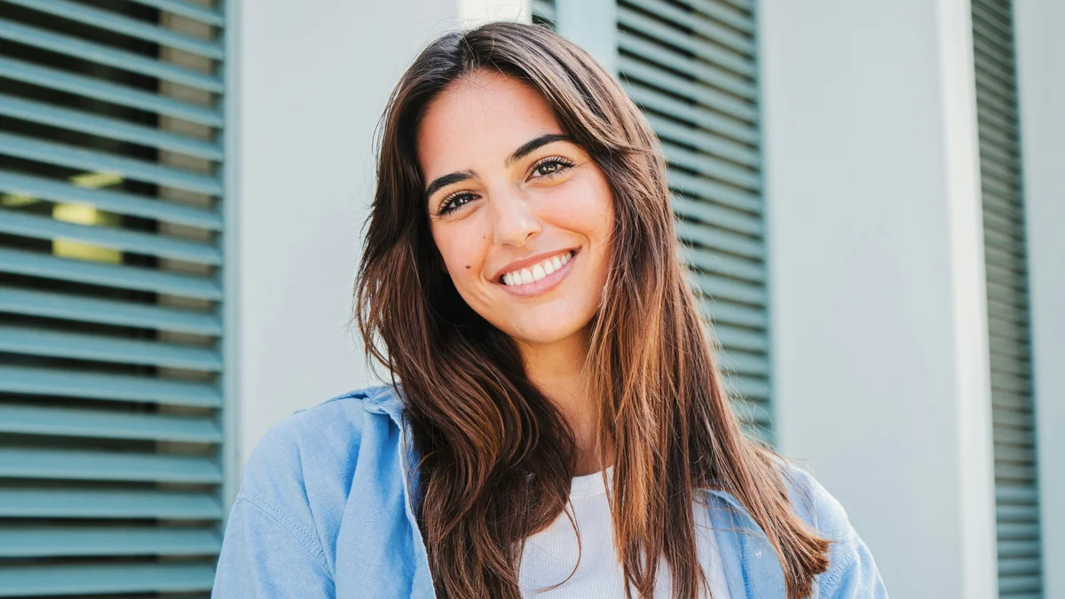 Une femme aux longs cheveux bruns et aux sourcils bien définis se tient à l'extérieur, souriante, devant un bâtiment aux volets verts