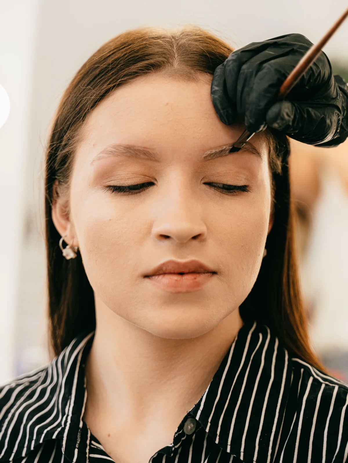 Une cliente assise en institut, les yeux fermés, pendant qu'une esthéticienne applique soigneusement la teinture avec un pinceau fin