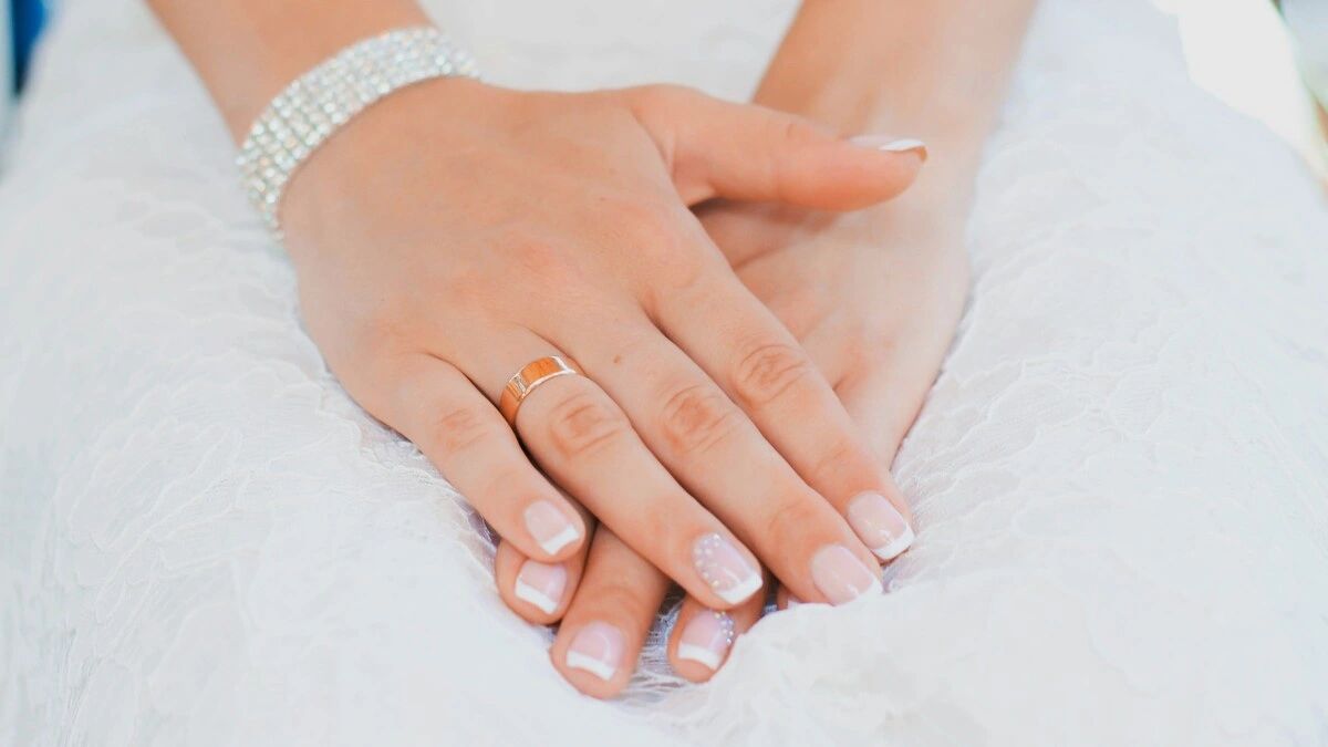 Les mains d'une femme reposent sur ses genoux ; elle porte une robe de mariée en dentelle et une jolie French manucure dans une association classique de nude et de blanc sur des ongles courts carrés-ovales, avec un délicat ornement sur les annulaires