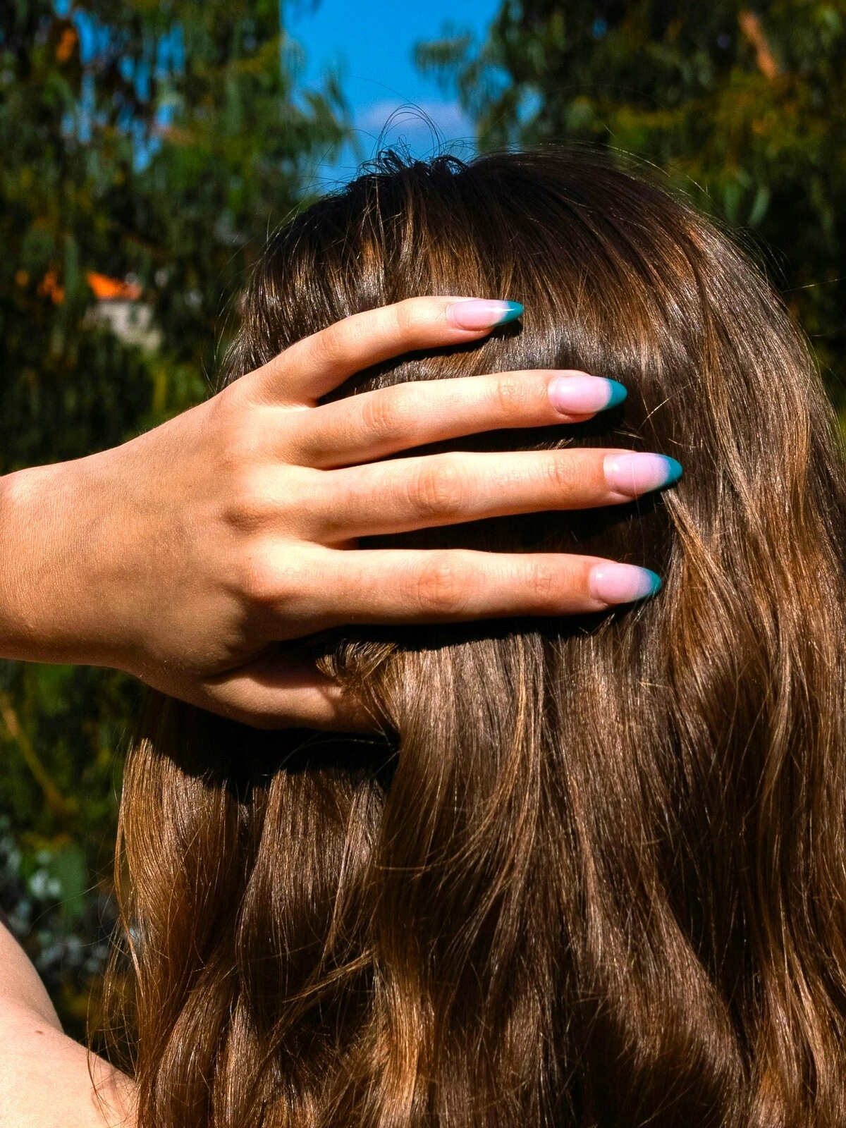 La main d'une femme posée à l'arrière de sa tête, arborant une French manucure avec une base rose pâle et des pointes bleu canard sur des ongles en amande