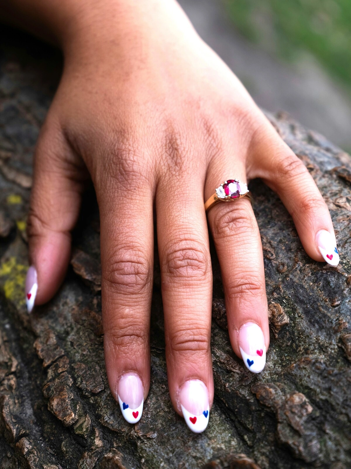 La main d'une femme posée sur une branche, arborant une French manucure sur des ongles ovales ornés de petits cœurs rouges et bleus