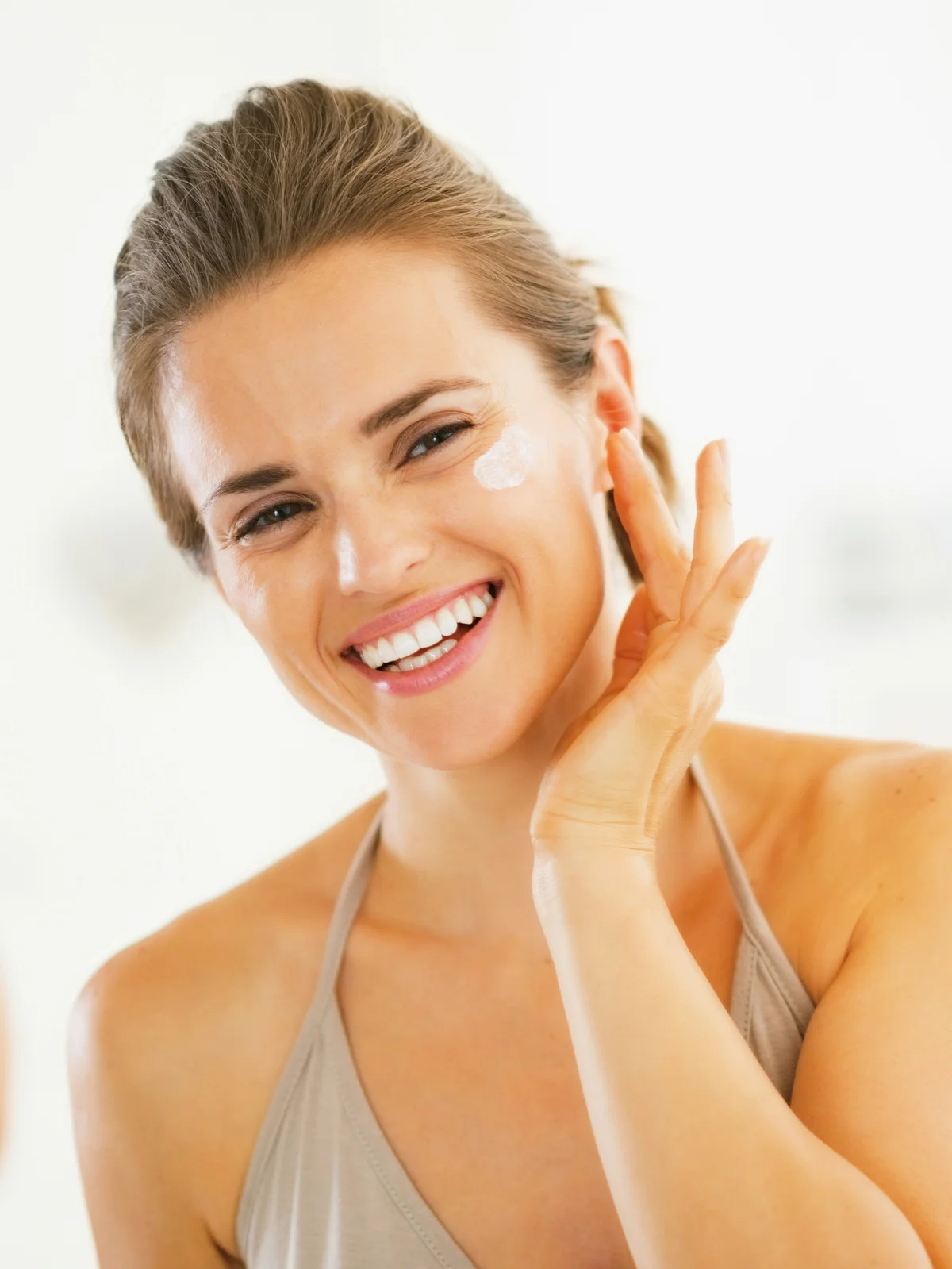 Gros plan d'une femme souriante, les cheveux attachés, appliquant une protection solaire sur son visage