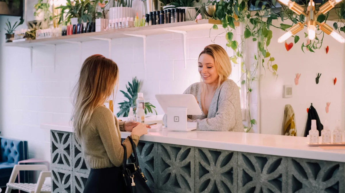 Une femme se tient au comptoir d'un institut pendant qu'une réceptionniste réserve son prochain soin ; l'institut est soigné, avec un comptoir blanc, des étagères de produits bien rangées et des plantes et des fleurs en décoration