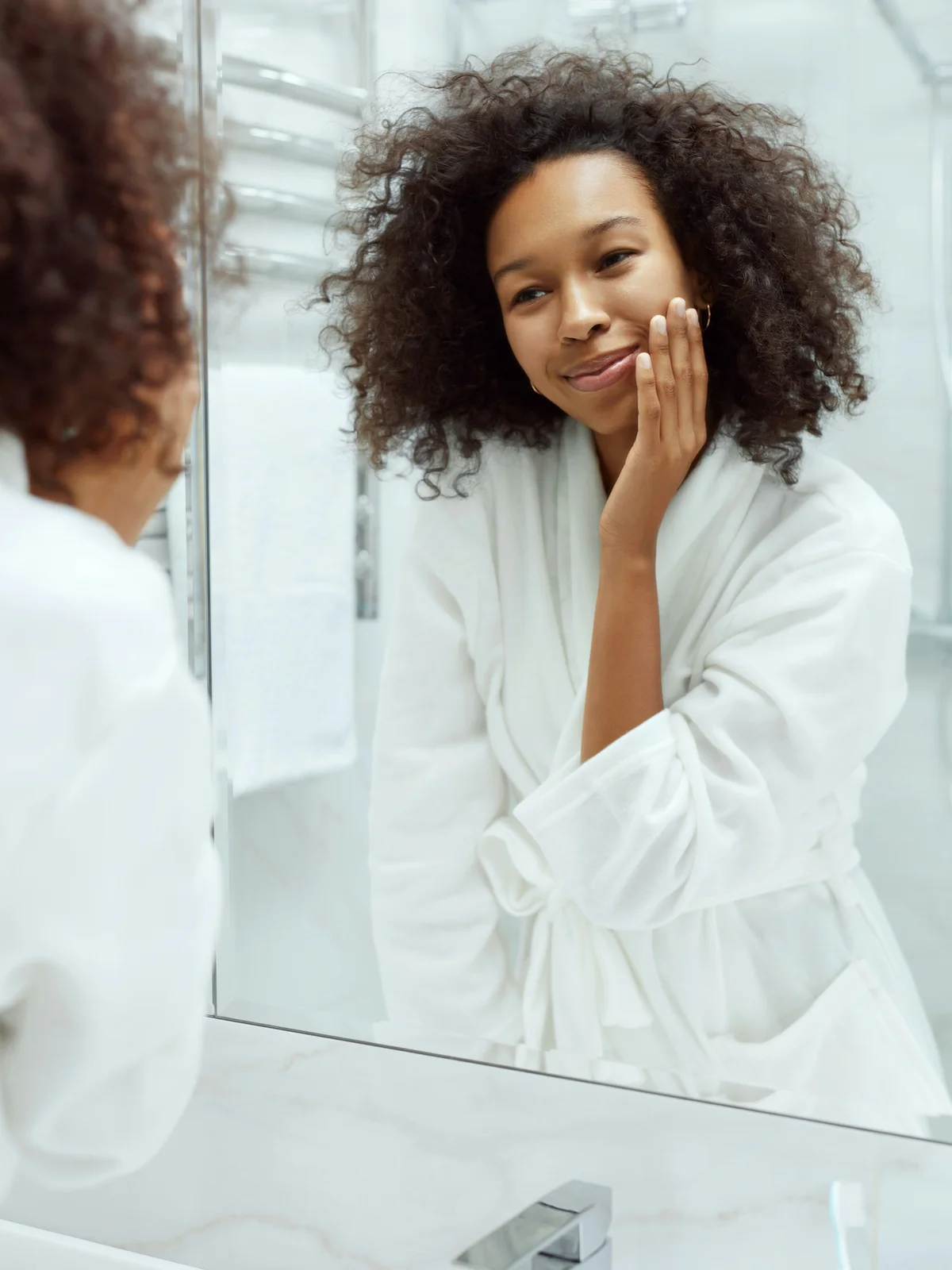 Une femme en peignoir blanc se regarde dans le miroir et effleure son visage, révélant une peau lisse et nette