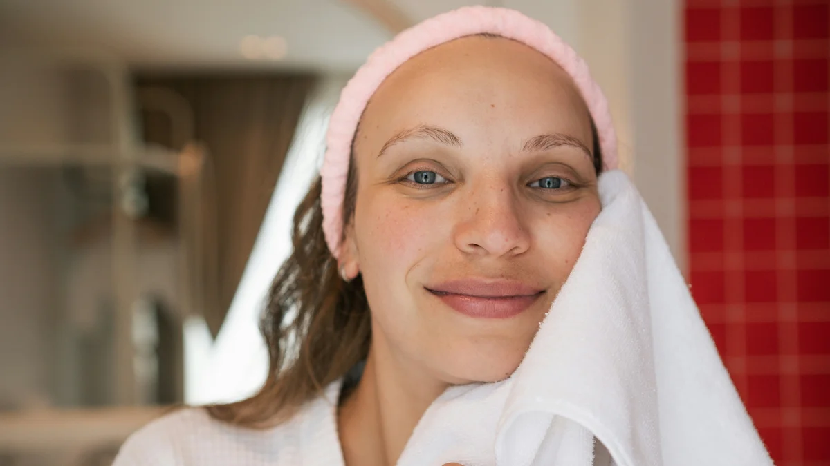 Une femme à la peau nette et lumineuse sourit face à l'objectif, un bandeau rose sur la tête, et tamponne son visage propre avec une serviette blanche