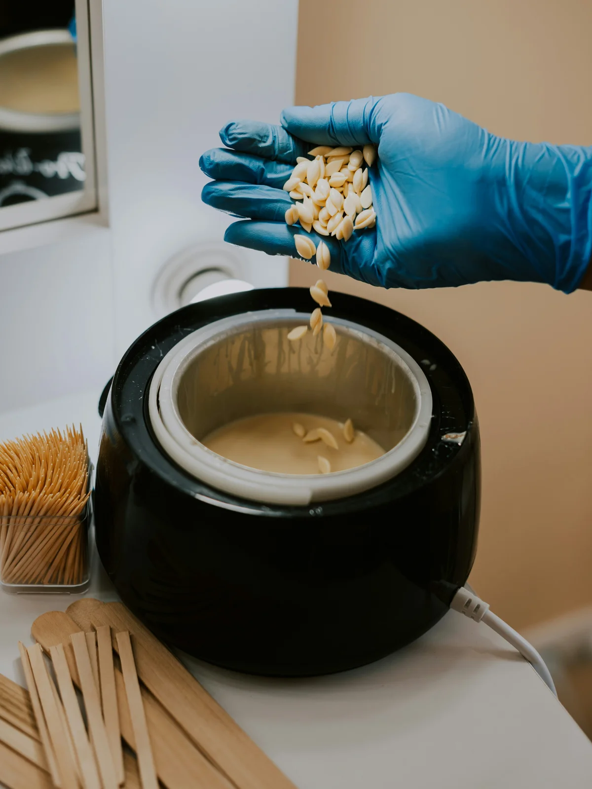 Gros plan sur la main gantée d’une esthéticienne mélangeant des perles de cire chaude dans un pot, prête pour un rendez-vous