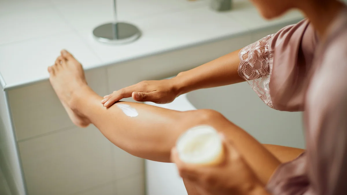 Prise en vue de dos d’une femme en peignoir en soie lilas appliquant une crème hydratante sur sa jambe, posée sur le rebord dans sa salle de bain