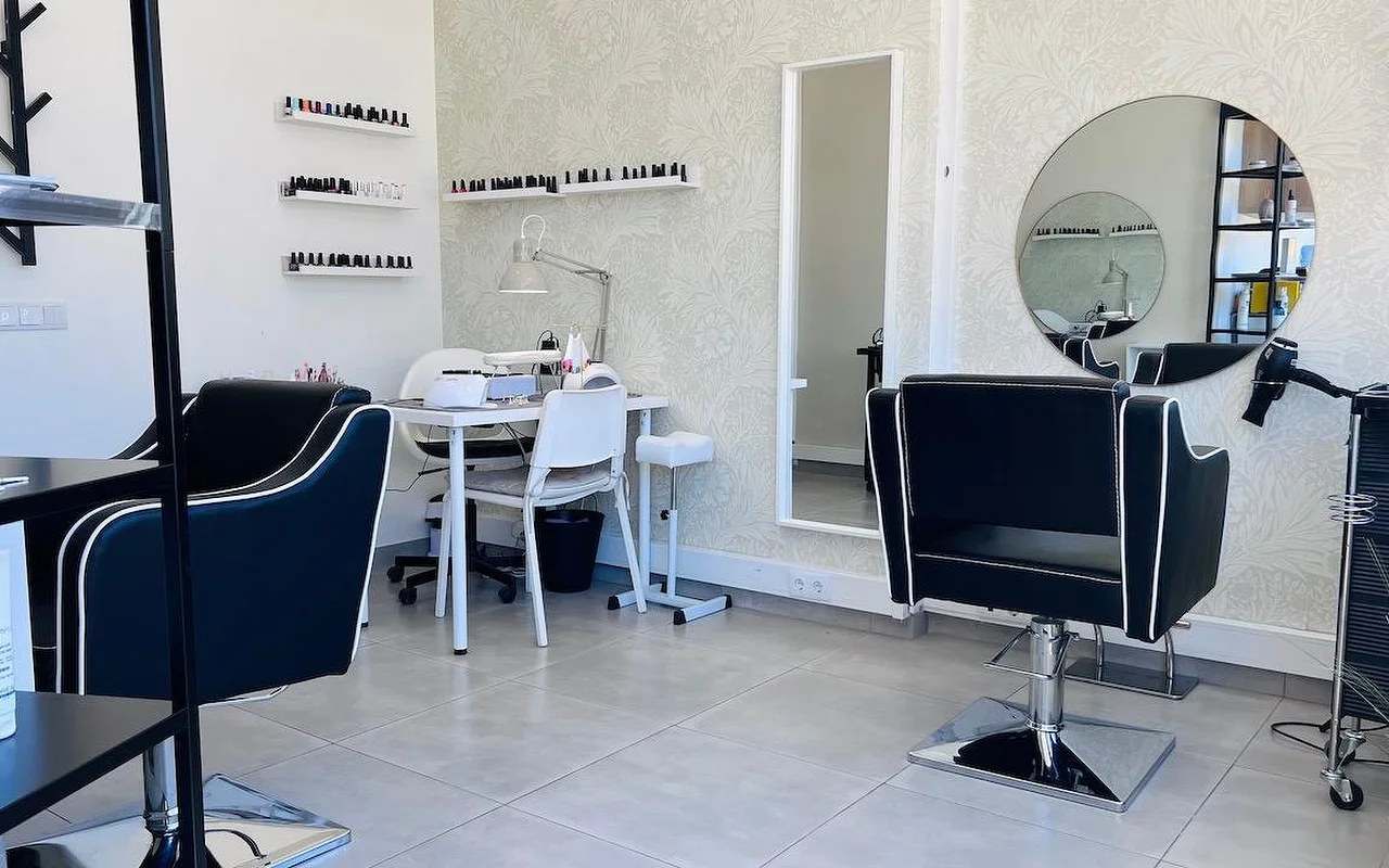 Un salon compact de coiffure et d'ongles avec des fauteuils de coiffage noirs soulignés d'un passepoil blanc, un miroir rond et un grand miroir sur un papier peint gris à motifs floraux, et des étagères murales alignant des vernis à ongles.