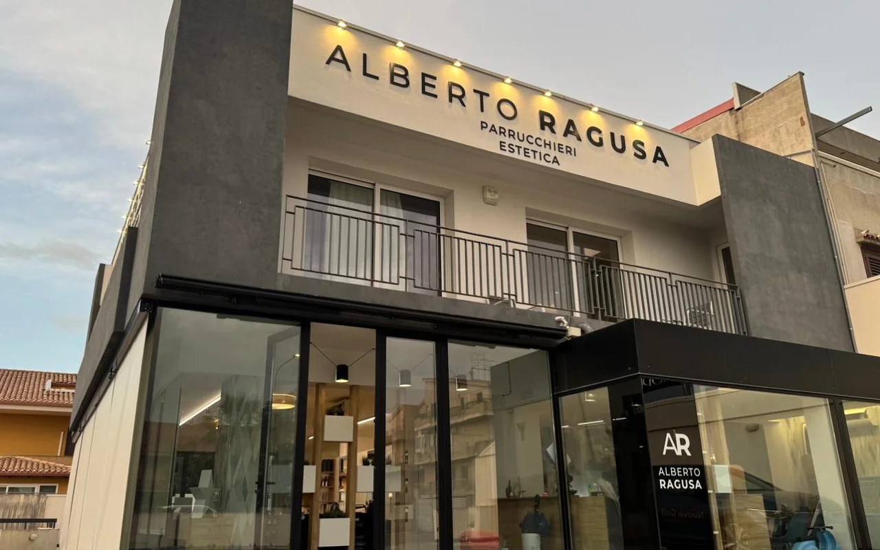 L'extérieur d'Alberto Ragusa au crépuscule, un salon sur deux niveaux avec une grande enseigne lumineuse indiquant « Alberto Ragusa Parrucchieri Estetica » au-dessus de baies vitrées du sol au plafond, une lumière chaude rayonnant de l'intérieur.