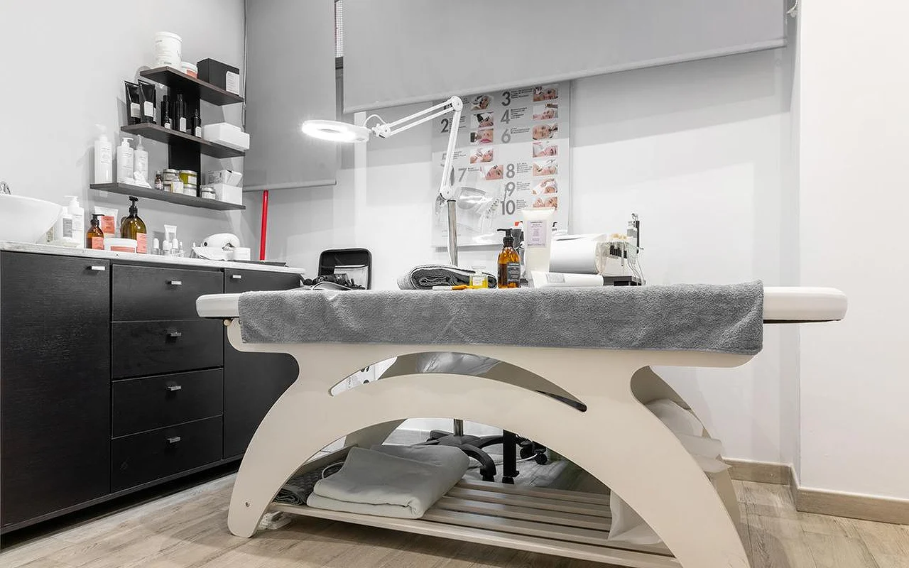 Une cabine d'esthétique compacte chez Atelier Beauty and the Lab, avec une table de soin crème, une lampe-loupe réglable, des étagères murales sombres garnies de produits de soin, et un schéma numéroté des étapes du soin au mur.