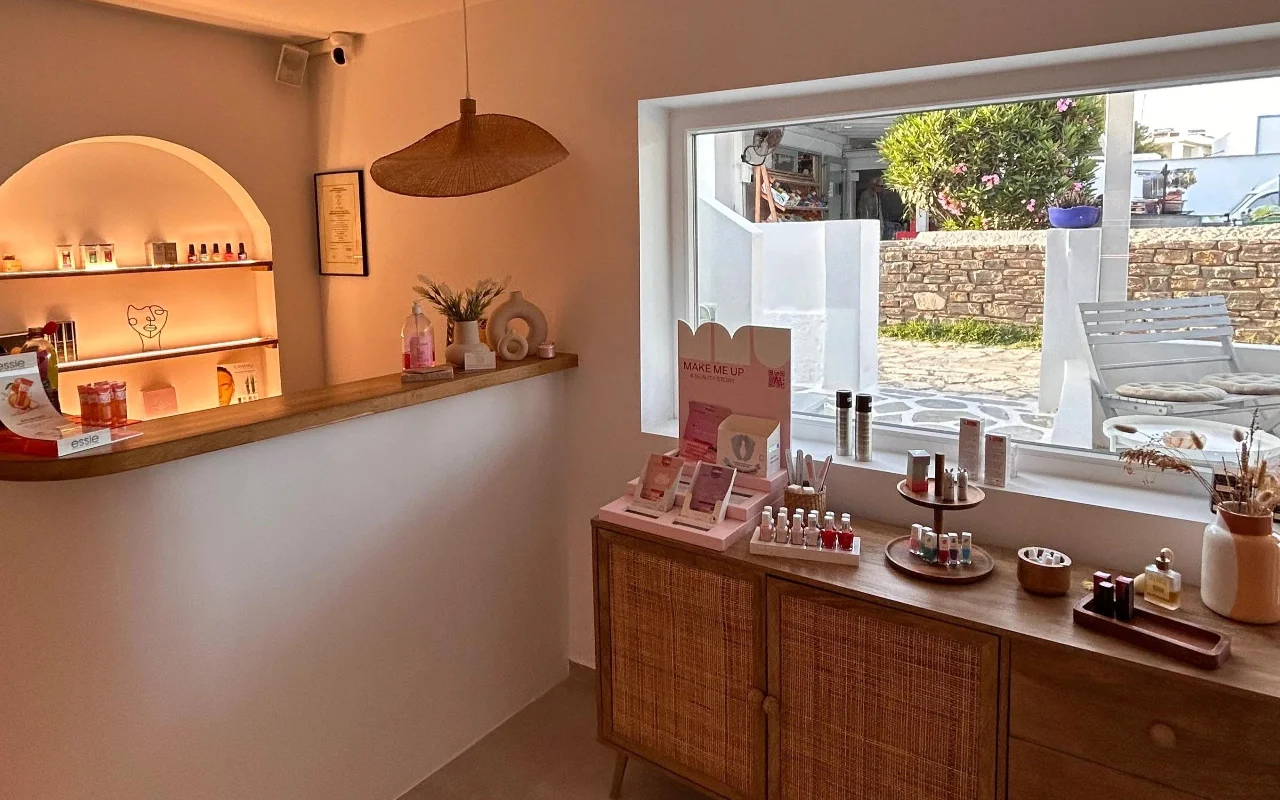 L'intérieur de BeautiQ Esthetics and More à Paros, avec une niche voûtée délicatement éclairée remplie de vernis à ongles, une suspension en rotin tressé et une grande fenêtre donnant sur un mur de pierre et un jardin ensoleillé.
