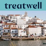 Un village de pêcheurs blanchi à la chaux de l'Algarve, aux maisons aux toits de terre cuite s'étageant à flanc de colline au-dessus d'un plan d'eau calme, avec de petits bateaux de pêche amarrés le long du quai, des palmiers et le clocher d'une église sous un ciel bleu limpide.