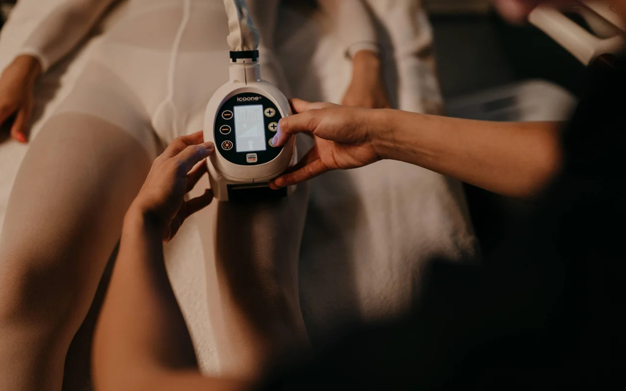 Un soin de remodelage du corps chez Beauty Room Romina Bongiovanni, avec une praticienne qui applique un appareil icoone sur la jambe d'une cliente en combinaison blanche de soin, dans une lumière chaude et tamisée.