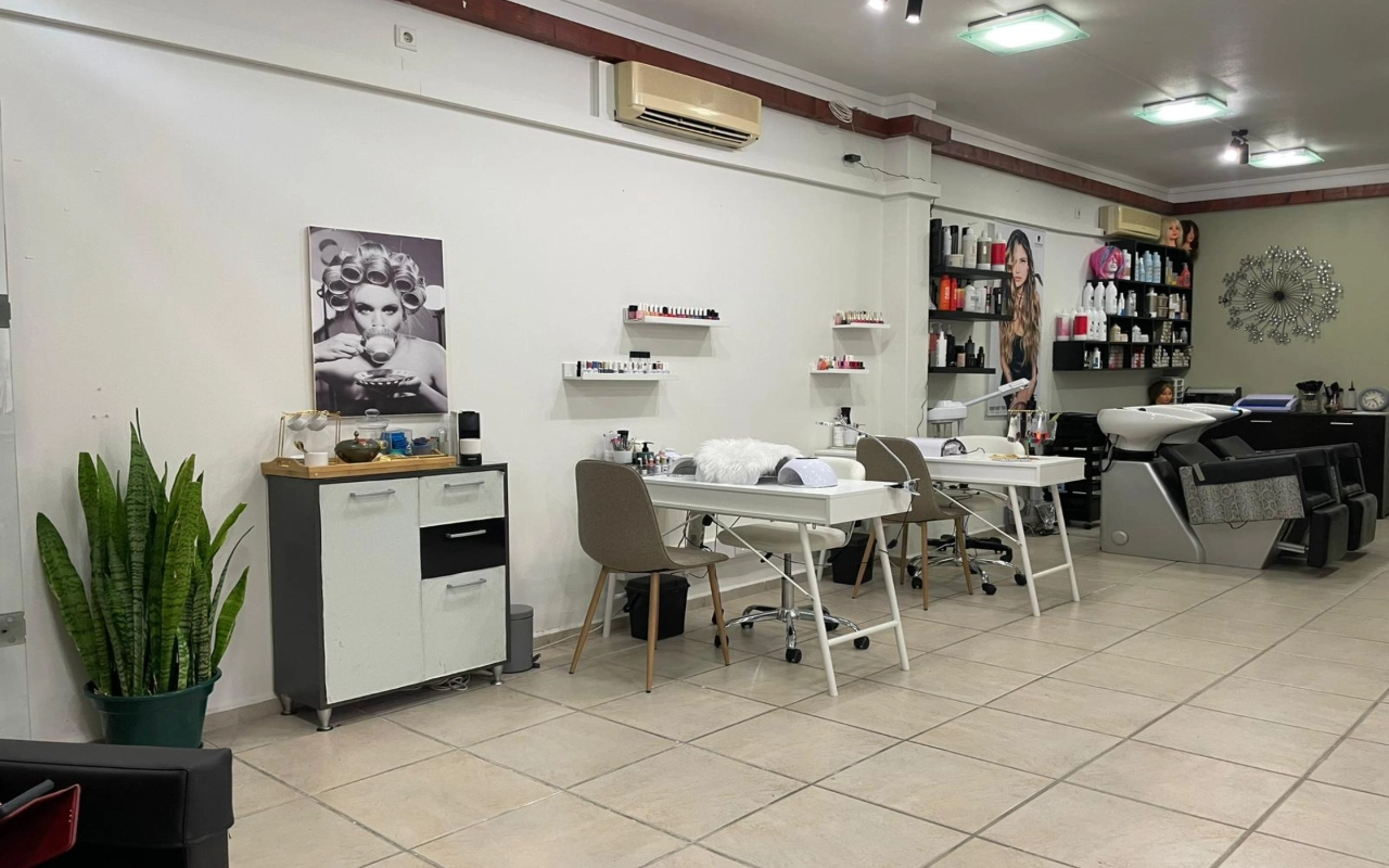 Un salon de coiffure et de beauté avec deux tables de manucure blanches et des chaises taupe, un bac de lavage et des étagères de produits le long du mur, une haute sansevière dans un coin, et une grande photographie en noir et blanc d'une femme aux bigoudis.