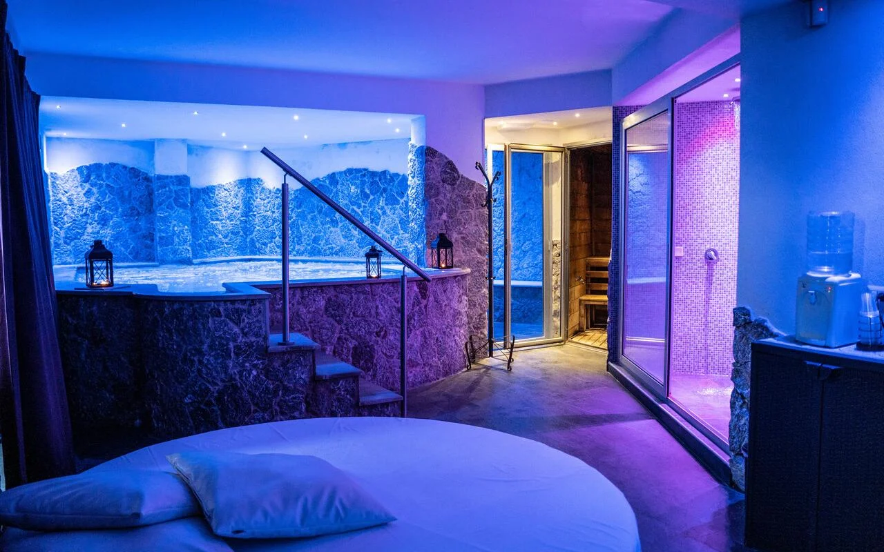 Une suite spa privée chez Bellezza e Benessere à Taormine, éclairée de bleu et de violet, avec un bain à remous encastré dans un habillage de pierre, un sauna en bois et, à l'arrière, un hammam en mosaïque, et un lit rond au premier plan.