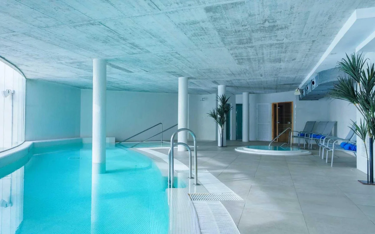 La piscine intérieure de spa de Benalmádena Beach, avec un bassin turquoise incurvé, un plafond en béton brut, un petit jacuzzi et des transats parmi de grands palmiers en pot.