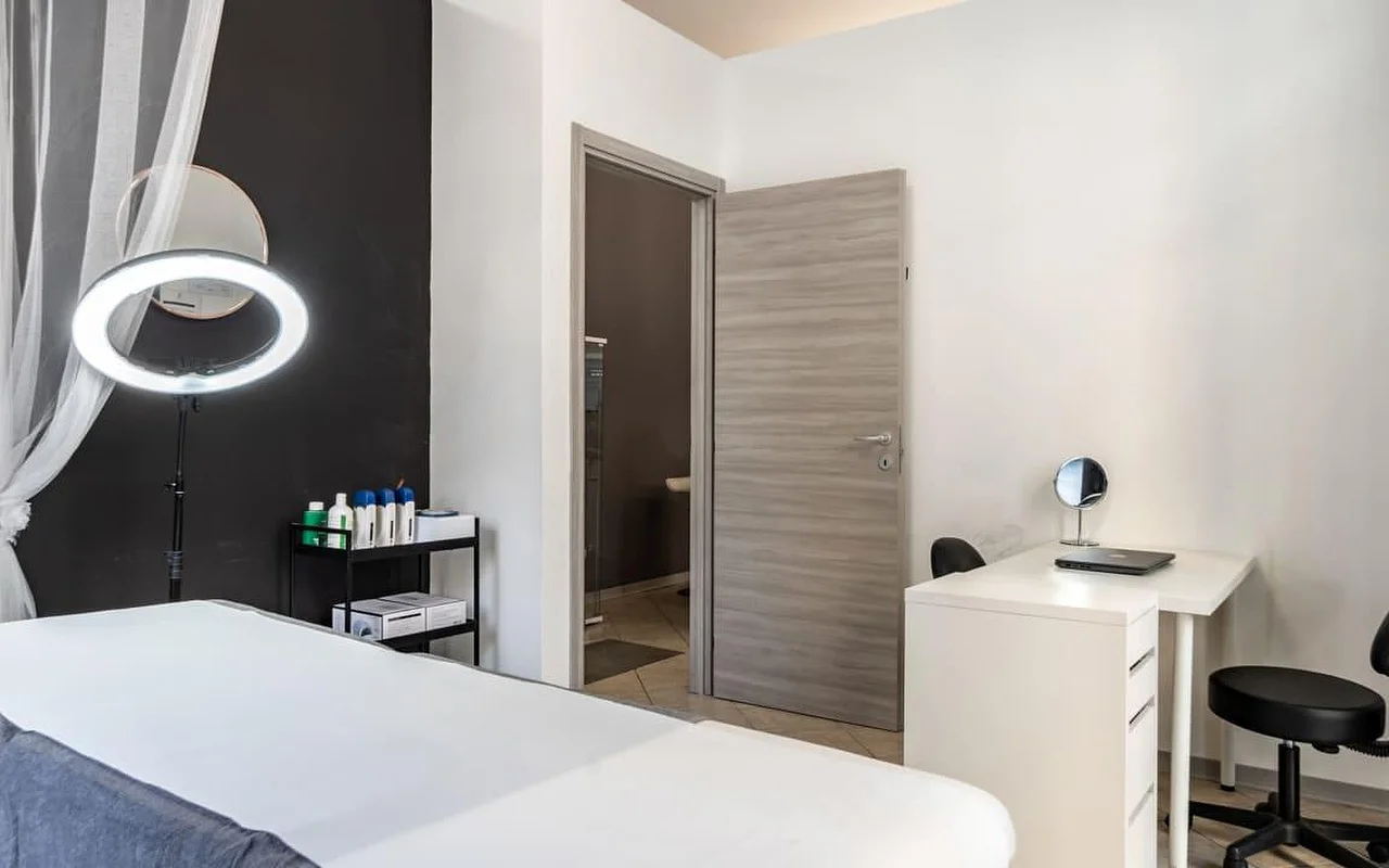 Une cabine de soin chez Coquette Wellness and Beauty, avec une table de soin blanche, une ring light éclairée sur pied contre un mur noir décoratif, des voilages à la fenêtre et un petit bureau blanc avec un miroir de courtoisie près d'une porte donnant sur une salle de bains privative.
