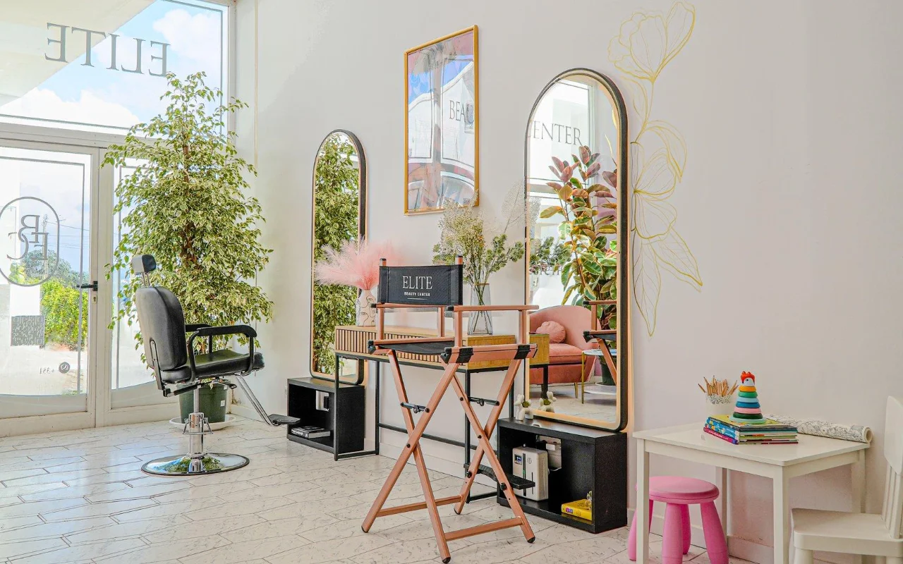Un salon de beauté lumineux et aéré, tout en blanc, avec des miroirs arqués aux cadres dorés, un fauteuil de coiffure noir et une chaise pliante de metteur en scène, de hautes plantes vertes, des touches de rose et un petit coin enfants près de la fenêtre.
