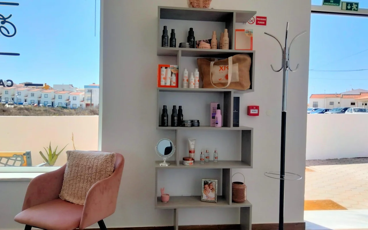 Un coin de salon près d'une grande fenêtre, avec une étagère grise en gradins garnie de produits capillaires et une photo encadrée, un fauteuil en velours rose poudré avec un coussin, et une vue sur de basses maisons blanchies à la chaux sous un ciel bleu.