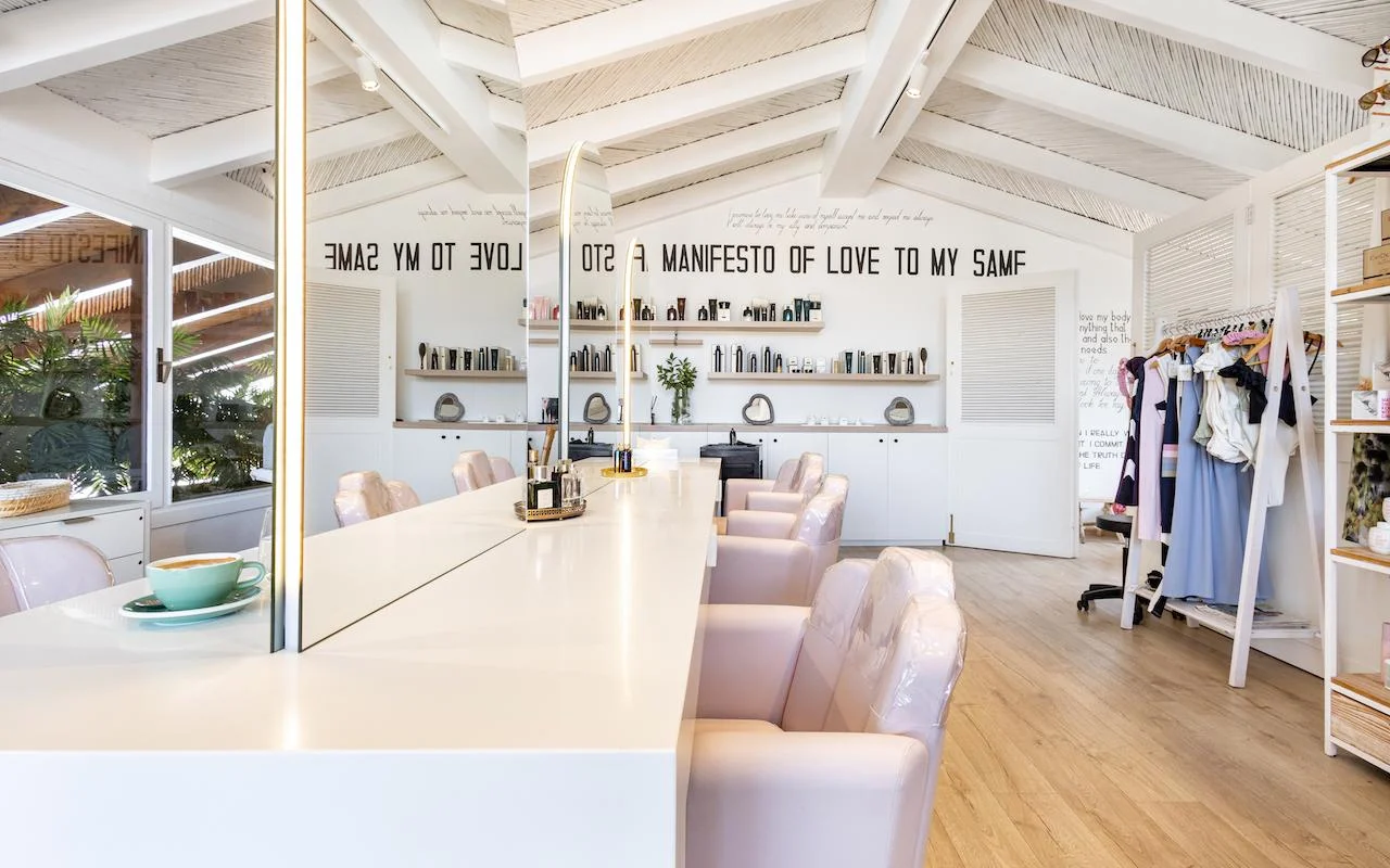 L'intérieur lumineux de Milk & Roses à Puente Romano, Marbella, avec un comptoir de coiffage blanc, des fauteuils rose pâle, un plafond à poutres et des étagères de produits le long du mur.