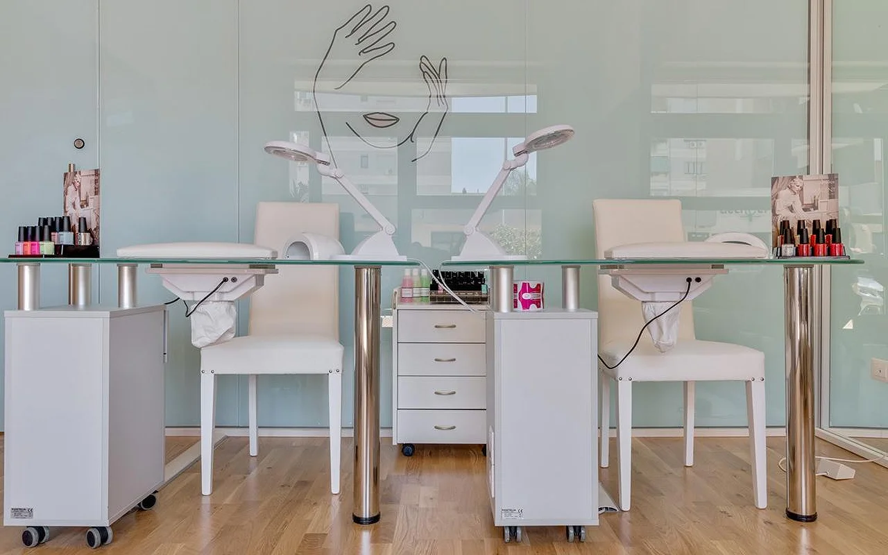 Un studio d'ongles chez Perfetta Urban Beauty, avec deux tables de manucure à plateau de verre et des chaises blanches, des lampes de travail, des présentoirs de vernis colorés, et une paroi de verre vert menthe portant le dessin au trait d'un visage et de deux mains.