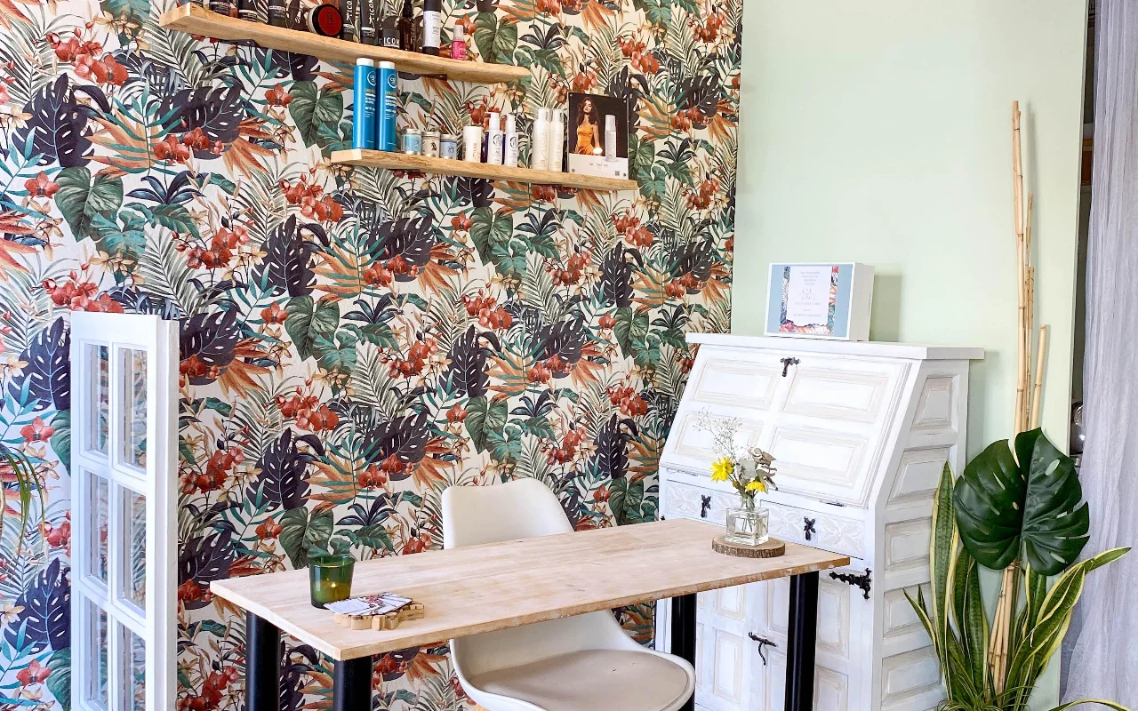 Un poste de coiffage chez Shangara Lora Organics à Marbella, avec un papier peint tropical audacieux, une table en bois, un ancien secrétaire blanc et des soins capillaires bio sur des étagères flottantes.