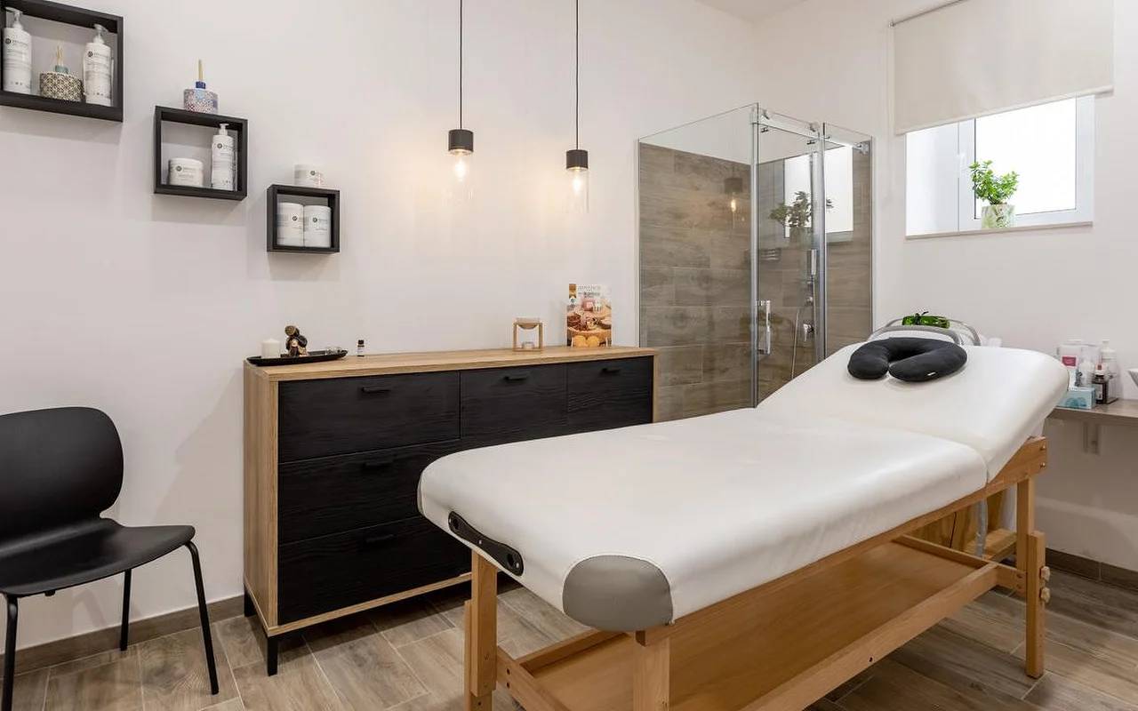 Une salle de massage chez Thea Estetica, avec une table de massage en bois surmontée d'un appui-visage noir, une commode noir et bois, deux suspensions, des étagères cube garnies de produits, et une cabine de douche en verre au carrelage effet bois.