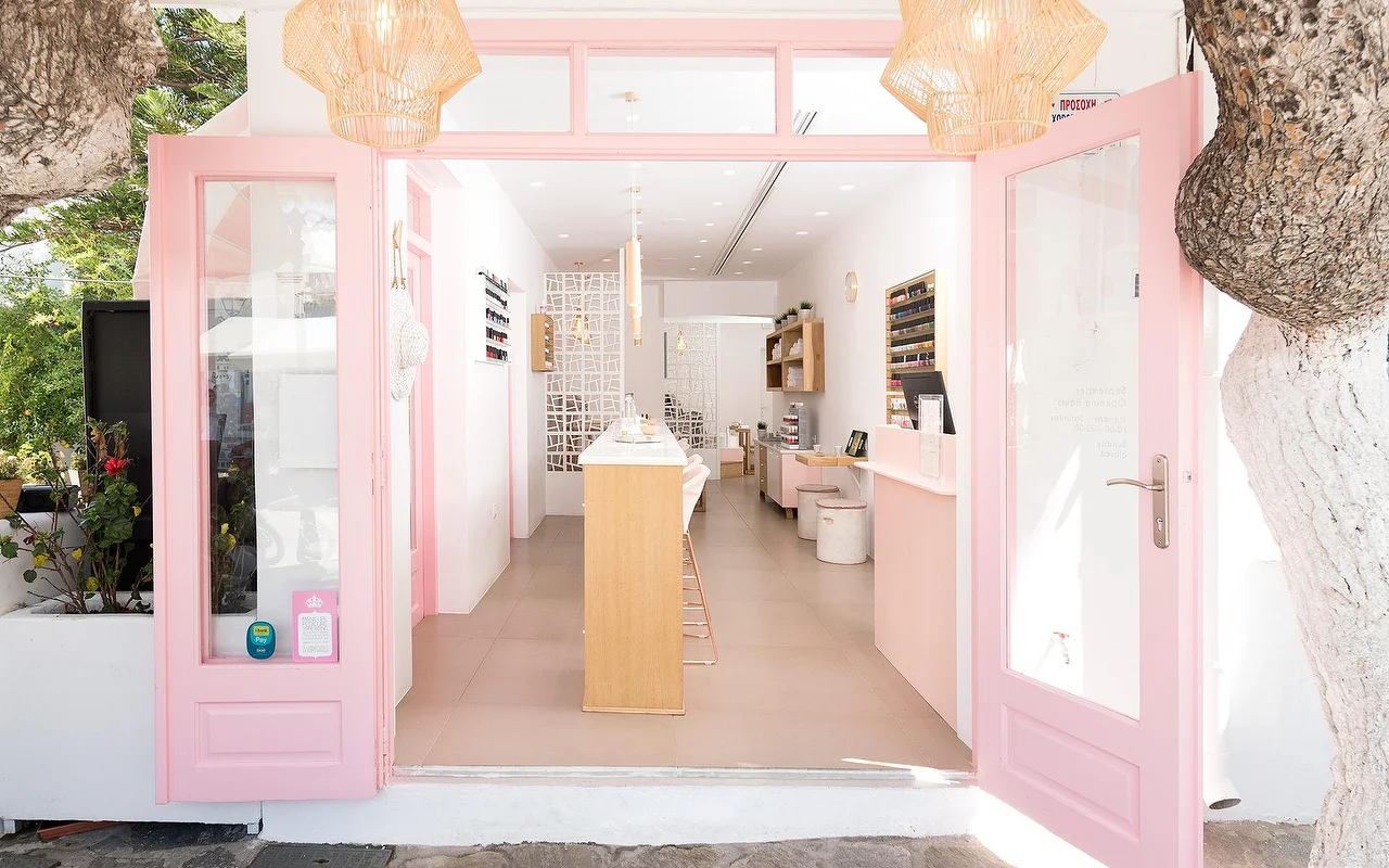 L'entrée encadrée de rose de Vivian Nail Boutique à Naoussa, Paros, portes ouvertes, suspensions tressées et comptoir en bois, dans un bâtiment cycladique blanchi à la chaux, à côté d'un arbre.