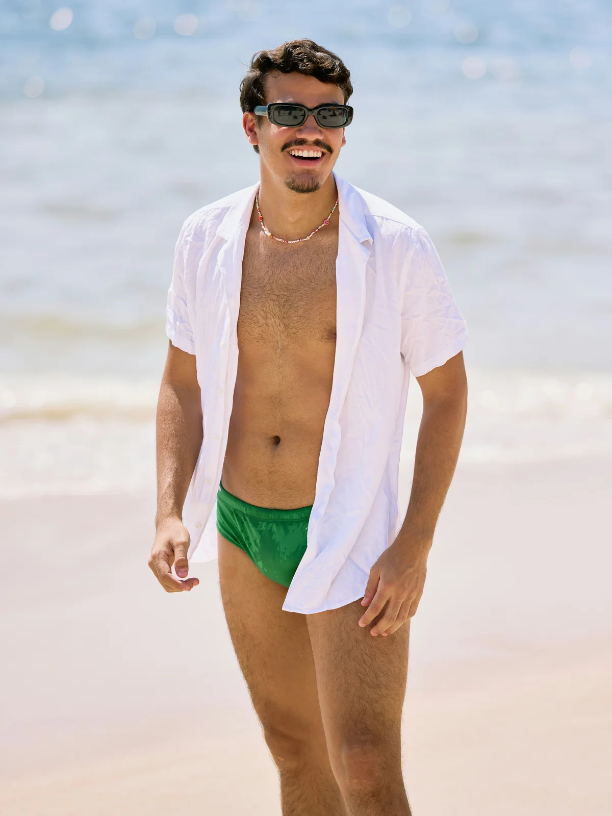 Un homme souriant debout sur une plage par une journée ensoleillée ; il porte un maillot de bain vert, une chemise blanche ouverte et des lunettes de soleil noires